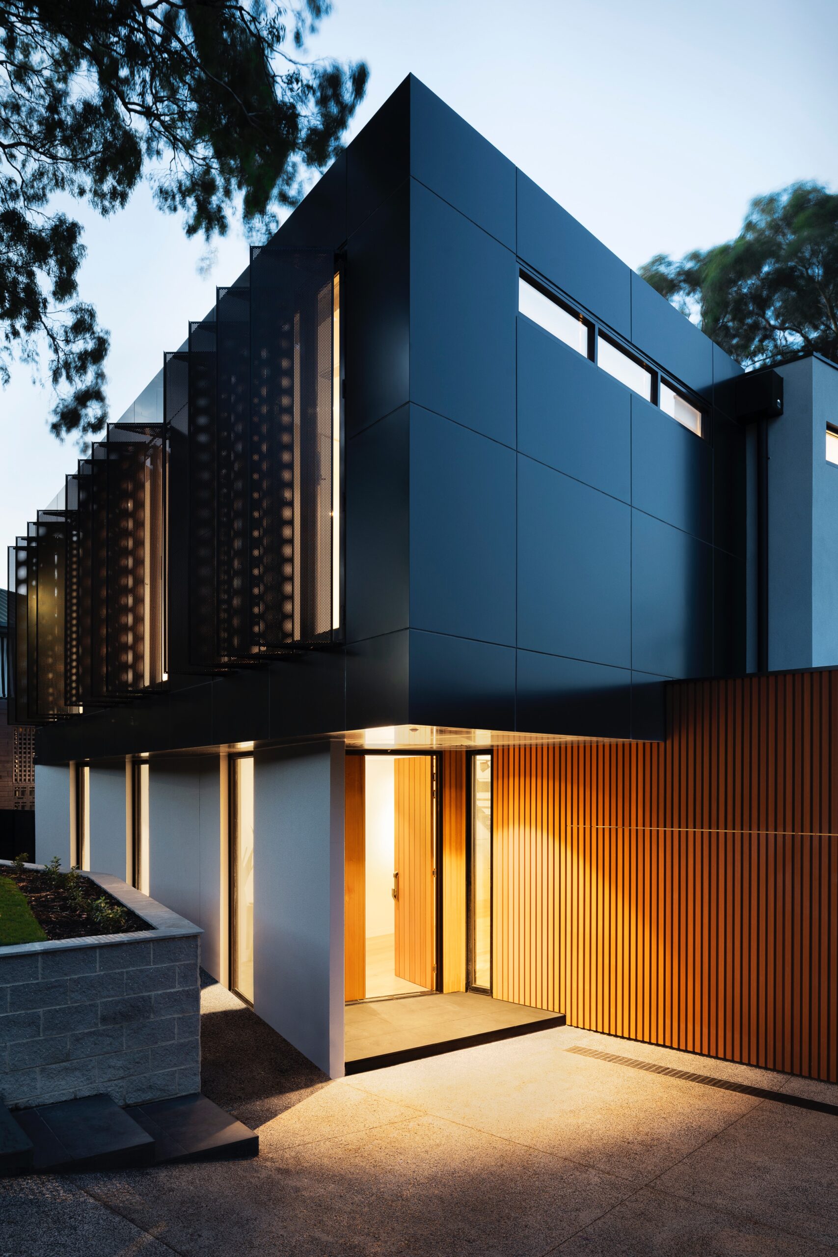 Modern home exterior with sleek black panels, wooden accents, and ambient lighting at dusk, surrounded by natural greenery.